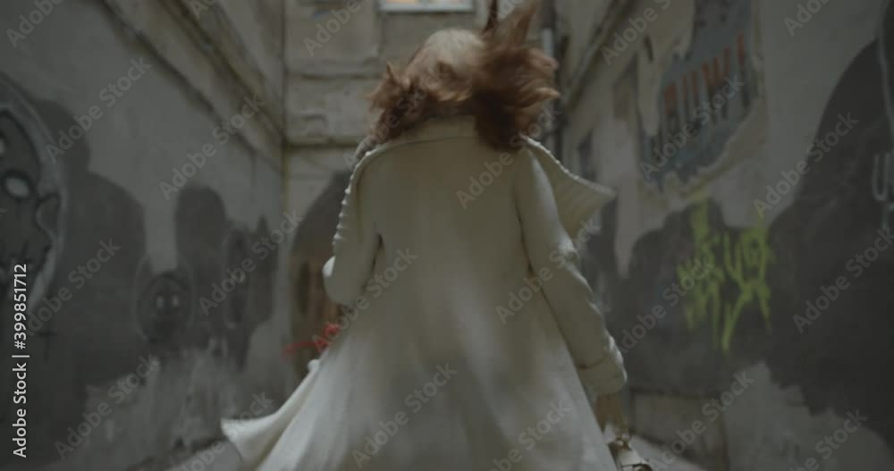 The young blonde woman in white coat is running away along the narrow old street. Rear close-up view.