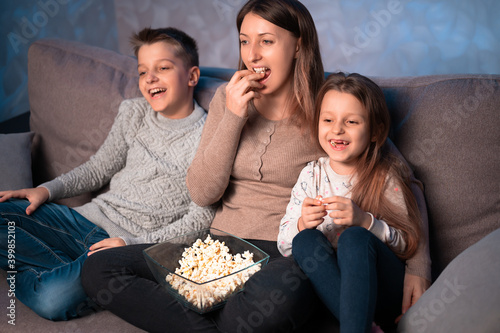 mom and kids sit on the couch in the evening  eat popcorn  watch TV and laugh.