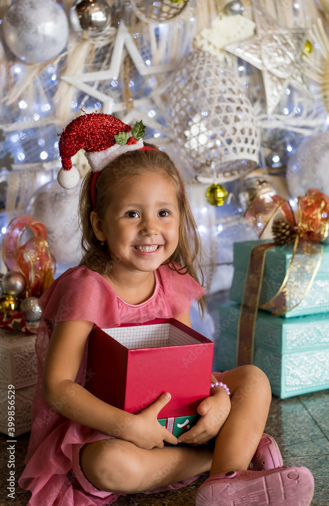 Adorable little girl wearing Santa hat sitting on the floor near Christmas tree and opening presents and gift box. Merry Christmas and happy holidays