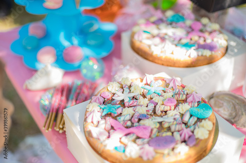 Mermaid Candy Bar. A beautiful cake decorated with mermaid tails.White shell plate full of chocolate candies with blue icing, cookies with shells, stars on the table with shining cloth. 