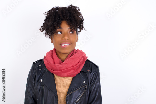 Young beautiful African American woman wearing biker jacket against white wall with thoughtful expression, looks away keeps hands down bitting his lip thinks about something pleasant.