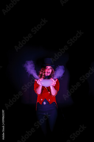 Female magician makes show with soap bubbles and smoke, an illusionist in theatrical clothes, on black wall. Woman actress in stage costume and top hat on her head. Concept of performance. Copy space