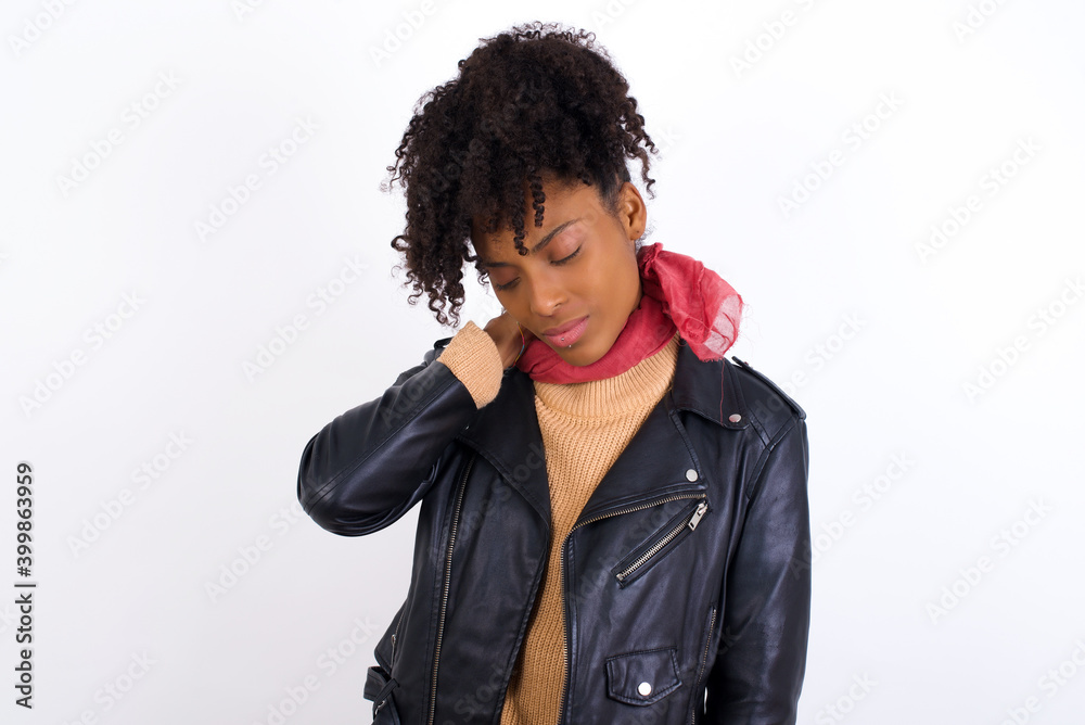 Young beautiful African American woman wearing biker jacket against white wall suffering from back and neck ache injury, touching neck with hand, muscular pain.