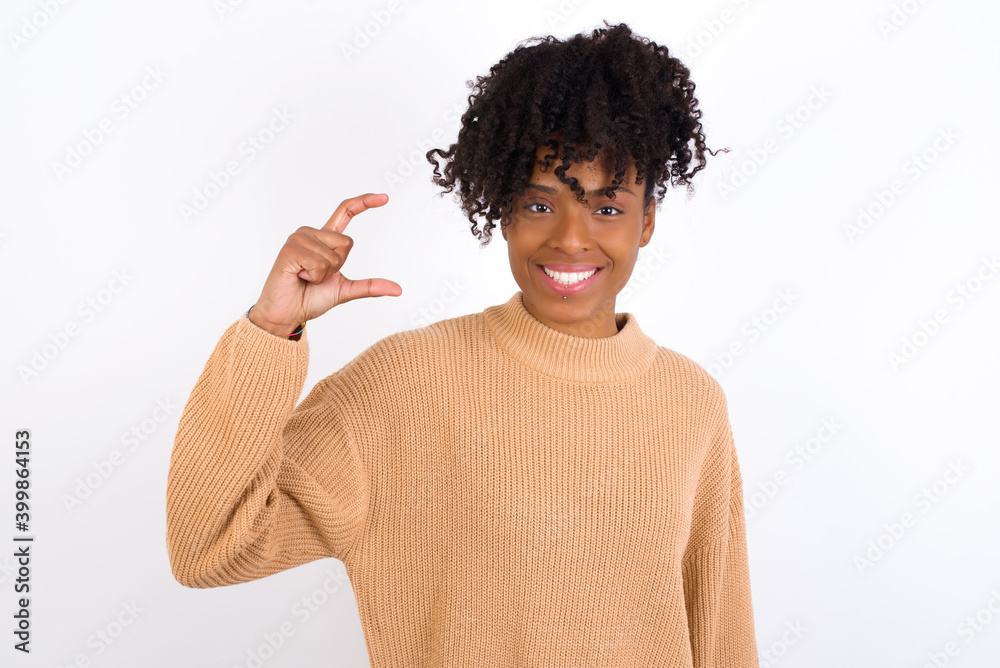 Young beautiful African American woman wearing knitted sweater against white wall, smiling and gesturing with hand small size, measure symbol.