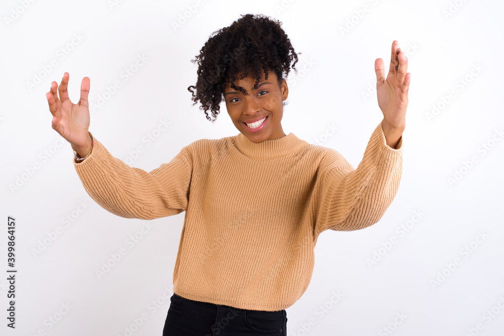 Cheerful Young beautiful African American woman wearing knitted sweater against white wall, making a welcome gesture raising arms over head. Cheerful Young beautiful African American woman wearing knitted sweater against white wall, making a welcome gesture raising arms over head.