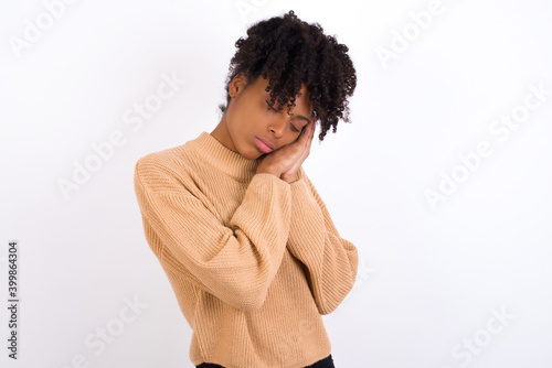 Relax and sleep time. Tired Young beautiful African American woman wearing knitted sweater against white wall with closed eyes leaning on palms making sleeping gesture.