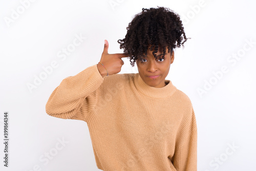 Unhappy Young beautiful African American woman wearing knitted sweater against white wall curves lips and makes suicide gesture, shoots in temple with hand, tries kill himself.