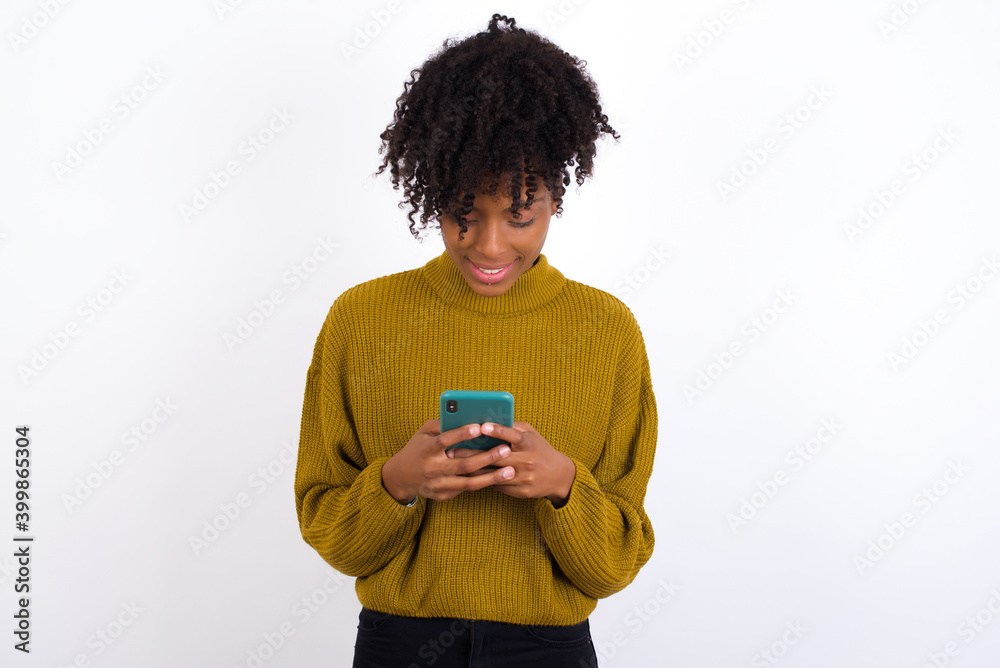 Smiling Young beautiful African American woman wearing knitted sweater using cell phone, messaging, being happy to text with friends, looking at smartphone. Modern technologies and communication. Smiling Young beautiful African American woman wearing knitted sweater using cell phone, messaging, being happy to text with friends, looking at smartphone. Modern technologies and communication.