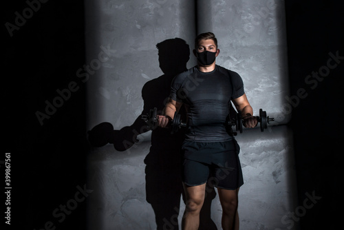 Male athlete wearing protective face mask and training with dumbbell in gym. Workout in gym after pandemic