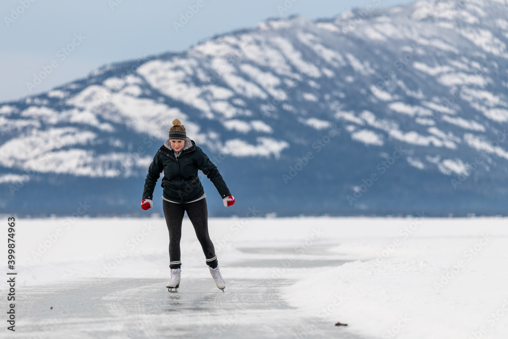 Woman skating on frozen lake in northern Canada with huge mountains in the background surrounded by snow, snow capped and glassy ice on lakes surface. 