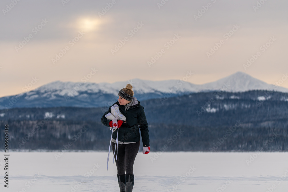 Woman walking towards camera while carrying skates with snow capped mountains in background, snowy landscape and Canada gloves. 