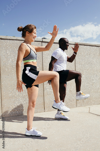 Red hair woman in grey top and black shorts doing exercises with jamaican fitness instructor in white t-shirt and black shorts on fresh air.