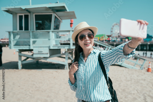 happy young female travel backpacker hold cellphone taking selfie with open lifeguard station. sunny day beach in Santa Monica los angeles USA. smiling girl tourist making self photo on mobile phone