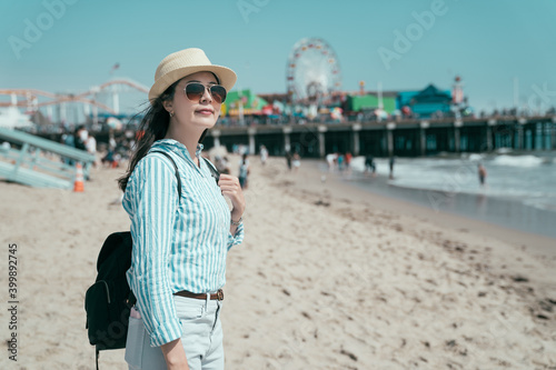 side happy smiling young girl traveler in straw hat and sunglasses carrying backpack. elegant female tourist standing sandy beach enjoy beautiful ocean view. bokeh of amusement park under blue sky.