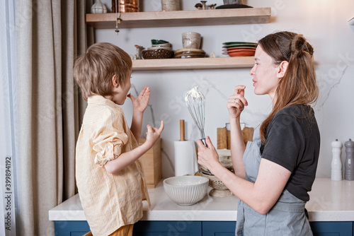 Family fun in the kitchen. Mother and son baking carrot cake together. Scandinavian kitchen interior. Mom holds a beater and plays with her son, they taste the cream