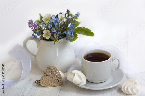 romantic breakfast: cup of coffee, meringues, a jug with forget-me-nots, wild strawberry flowers, white ribbon, carved wooden heart on a light background