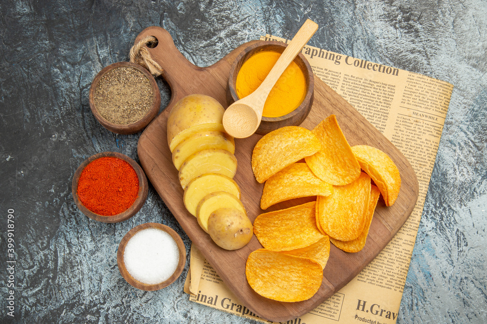 Above view of tasty homemade chips cut potato slices on wooden cutting board and different spices on newspaper on gray background Above view of tasty homemade chips cut potato slices on wooden cutting board and different spices on newspaper on gray background