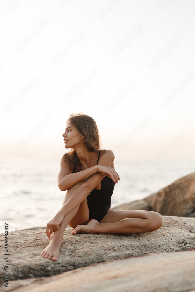 Beautiful woman in black swimsuit posing on the beach sitting on rocks, ocean view landscape