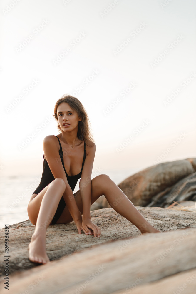 Beautiful woman in black swimsuit posing on the beach sitting on rocks, ocean view landscape Beautiful woman in black swimsuit posing on the beach sitting on rocks, ocean view landscape