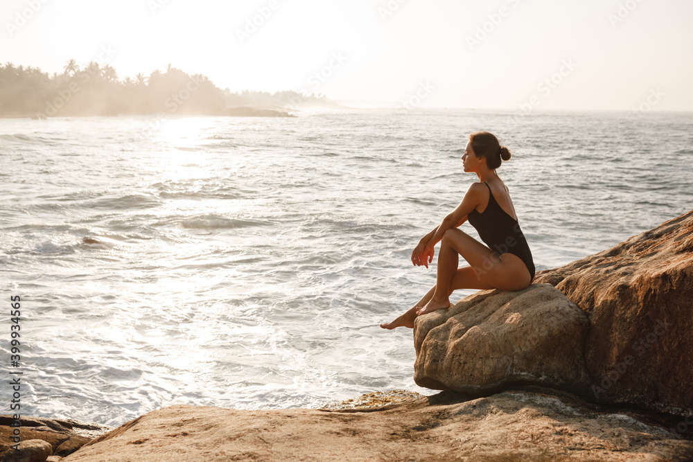 Beautiful woman in black swimsuit posing on the beach sitting on rocks, ocean view landscape