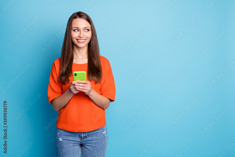 Photo of toothy beaming girl dressed casual jeans orange t-shirt holding hands arms device looking empty space isolated blue color background Photo of toothy beaming girl dressed casual jeans orange t-shirt holding hands arms device looking empty space isolated blue color background
