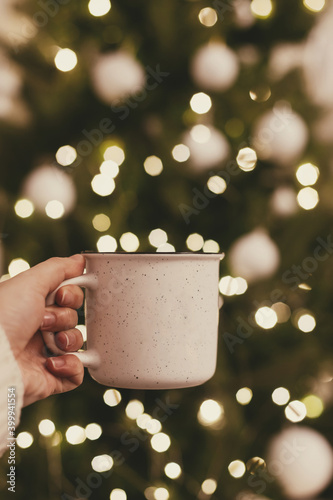 Hand holding festive cup of tea on background of Christmas tree lights bokeh. Merry Christmas and Happy Holidays!