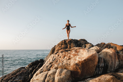 Woman model in black swimsuit is posing on the beach with big stones in background. Beautiful tanned girl. Concept bikini, swimwear
