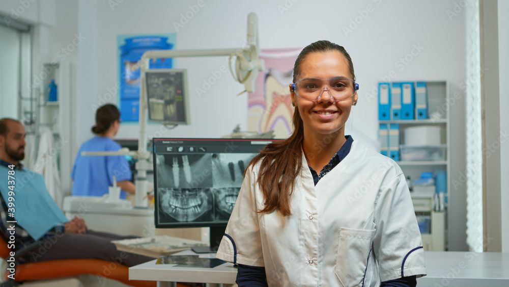 Stomatologist doctor smiling at camera in dental office while assistant preparing man for stomatological surgery in background. Woman dentist looking on webcam sitting on chair in stomatological room Stomatologist doctor smiling at camera in dental office while assistant preparing man for stomatological surgery in background. Woman dentist looking on webcam sitting on chair in stomatological room