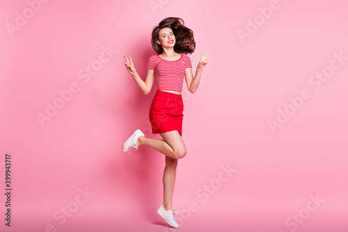 Full size photo of young beautiful smiling cheerful girl showing v-sign flying hair jump isolated on pink color background