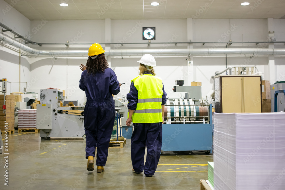 Back view of female factory workers walking and talking about work. Content experienced women working together at manufacturing plant and wearing protective uniform. Manufacture and occupation concept Back view of female factory workers walking and talking about work. Content experienced women working together at manufacturing plant and wearing protective uniform. Manufacture and occupation concept