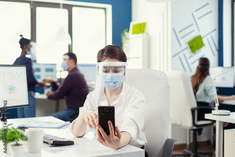 Manager using smartphone for online conference wearing face mask against covid-19 as safety precaution. Colleagues working respecting social distance during global pandemic with coronavirus.