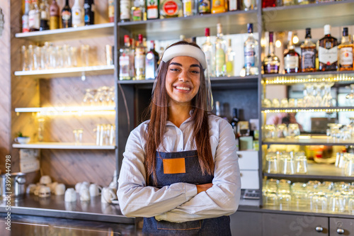 Portrait of a beautiful bartender standing at the counter smiling and looking at the camera while while wearing face shield due to covid-19, shelves full of bottles with alcohol on the background