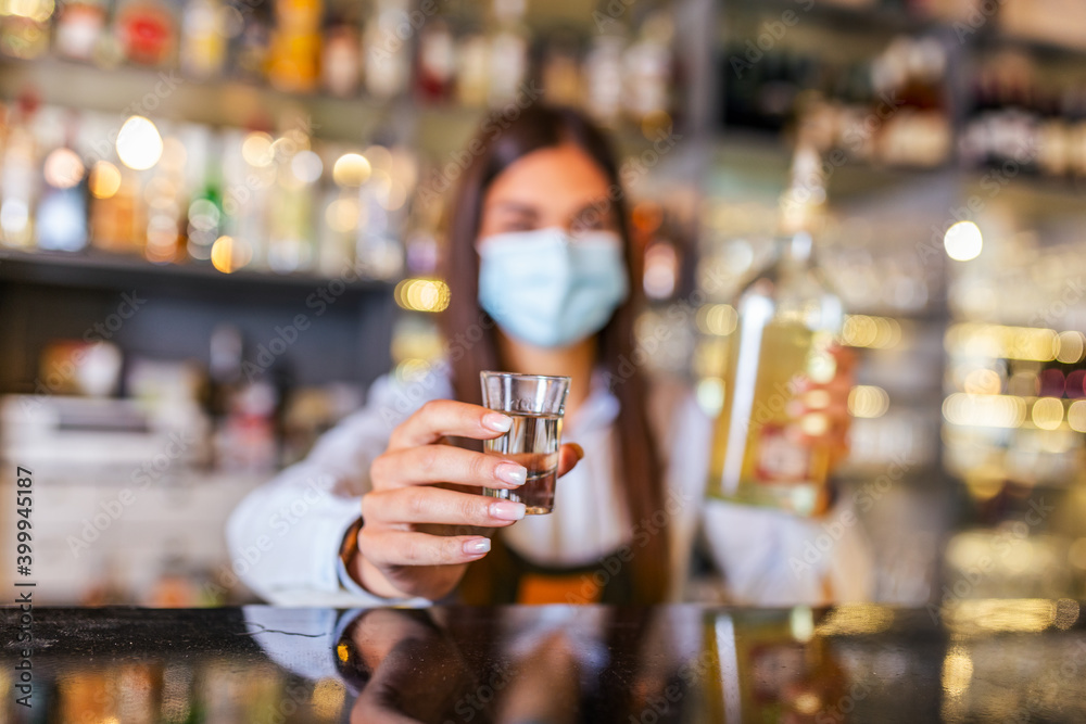 Beautiful female bartender with protective face mask pouring alcohol into shot glass during coronavirus pandemic, shelves full of bottles with alcohol on the background