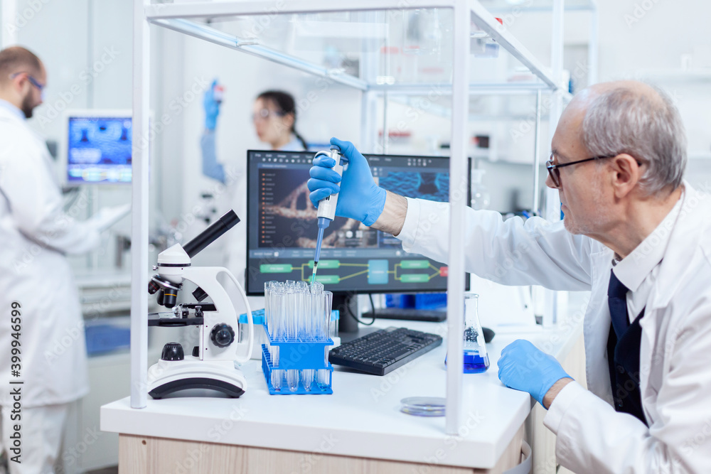 Experienced scientist working in busy laboratory at his workplace holding dropper dispense. Senior professional chemist using pippete with blue solution for microbiology tests. Experienced scientist working in busy laboratory at his workplace holding dropper dispense. Senior professional chemist using pippete with blue solution for microbiology tests.