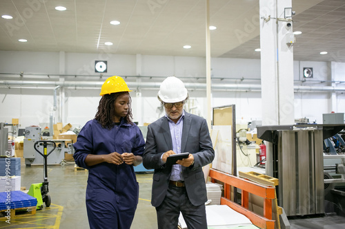 Caucasian manager discussing factory work with African worker. Beautiful pensive young woman working on plant and looking on tablet in supervisor hands. Manufacture and digital technology concept