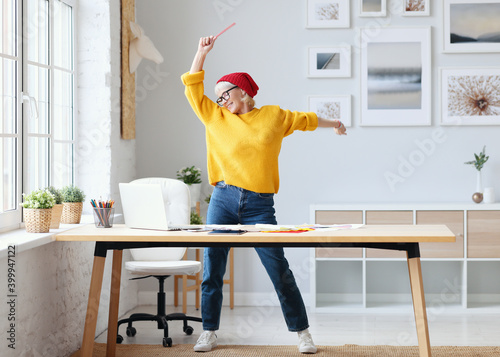 Photographie cheerful elderly woman freelancer creative designer in a red hat having fun and dancing in workplace