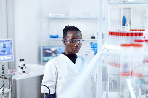 African scientist holding test tube with blue liquid for medical purpose. Black researcher in sterile laboratory conducting pharmacology experiment wearing coat.