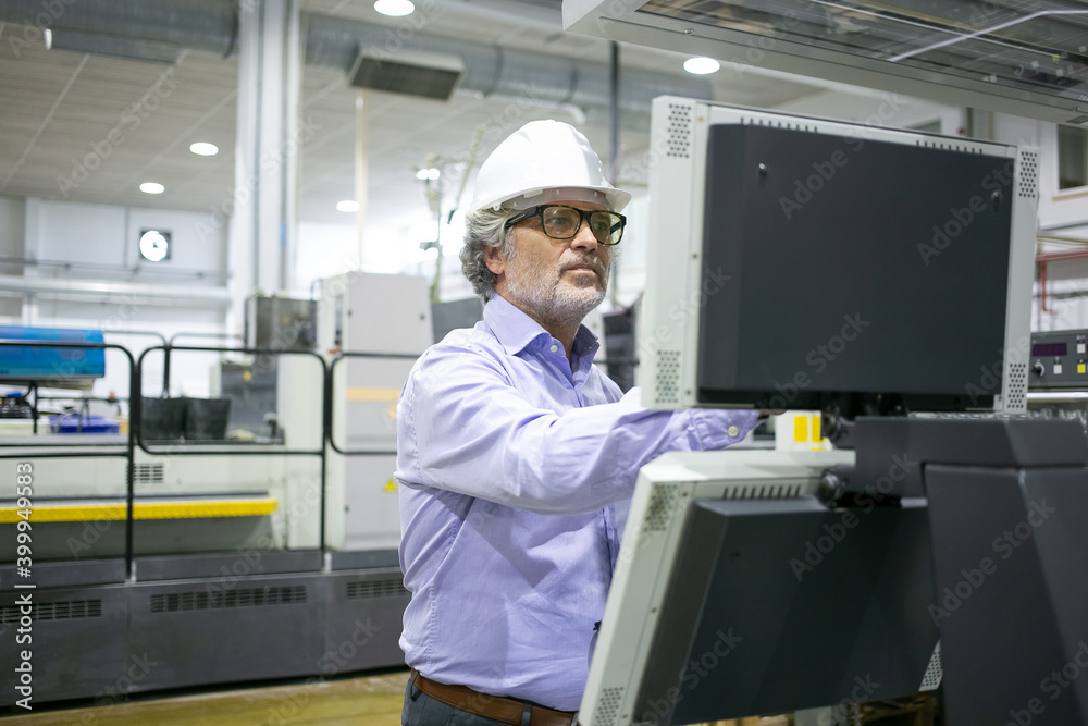 Serious male plant manager in hardhat and glasses operating industrial machine, pushing buttons on control panel. Copy space. Industrial technology or machinery concept Serious male plant manager in hardhat and glasses operating industrial machine, pushing buttons on control panel. Copy space. Industrial technology or machinery concept