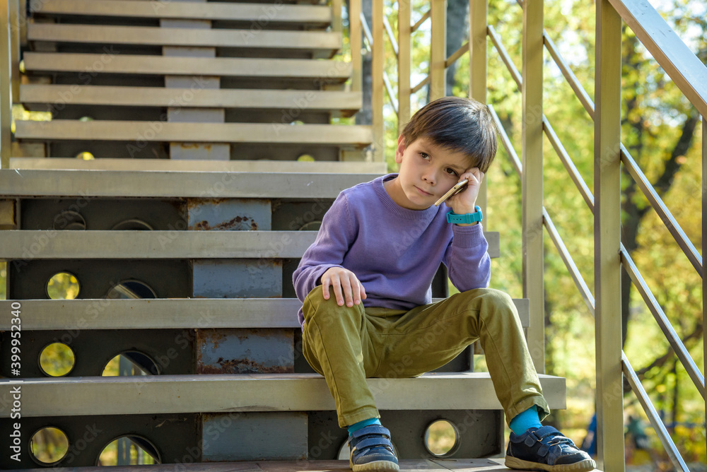 Closeup portrait, worried, sad young child, boy talking on phone to someone, looking unhappy, isolated outdoors background. Negative human emotions, facial expressions, feeling, reaction Communication Closeup portrait, worried, sad young child, boy talking on phone to someone, looking unhappy, isolated outdoors background. Negative human emotions, facial expressions, feeling, reaction Communication