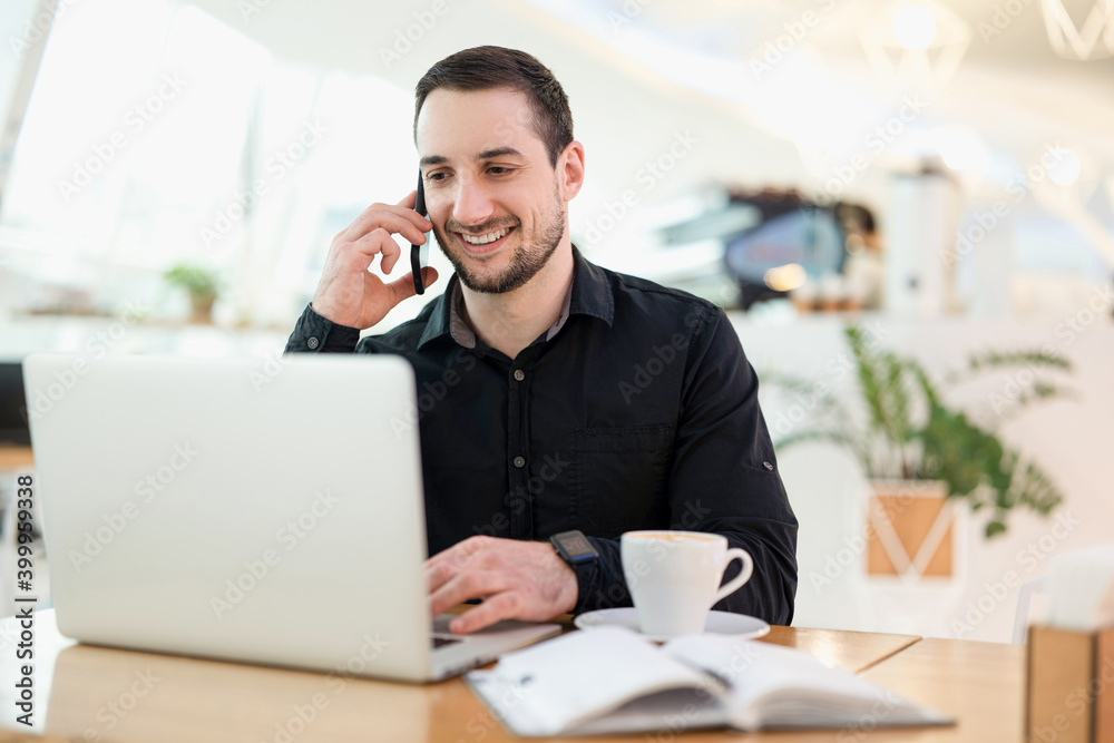 Succesful remote male worker. Young freelancer man happy with a phone call. Coffee shop on background. Male freelancer programmer working in a coffee shop. He is happy to speak with the client. Succesful remote male worker. Young freelancer man happy with a phone call. Coffee shop on background. Male freelancer programmer working in a coffee shop. He is happy to speak with the client.