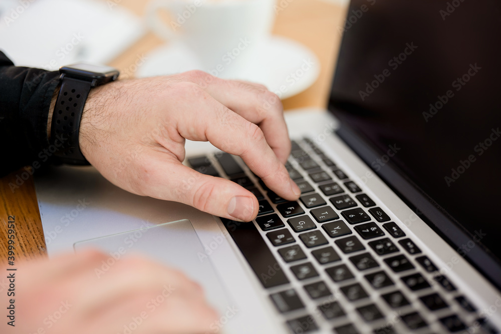 Male hands typing on laptop keyboard. Cut view. Modern gadgets. Close up. Gray laptop. Blurred cup of coffee on background. Man in black shirt. Male freelancer working from cafe.