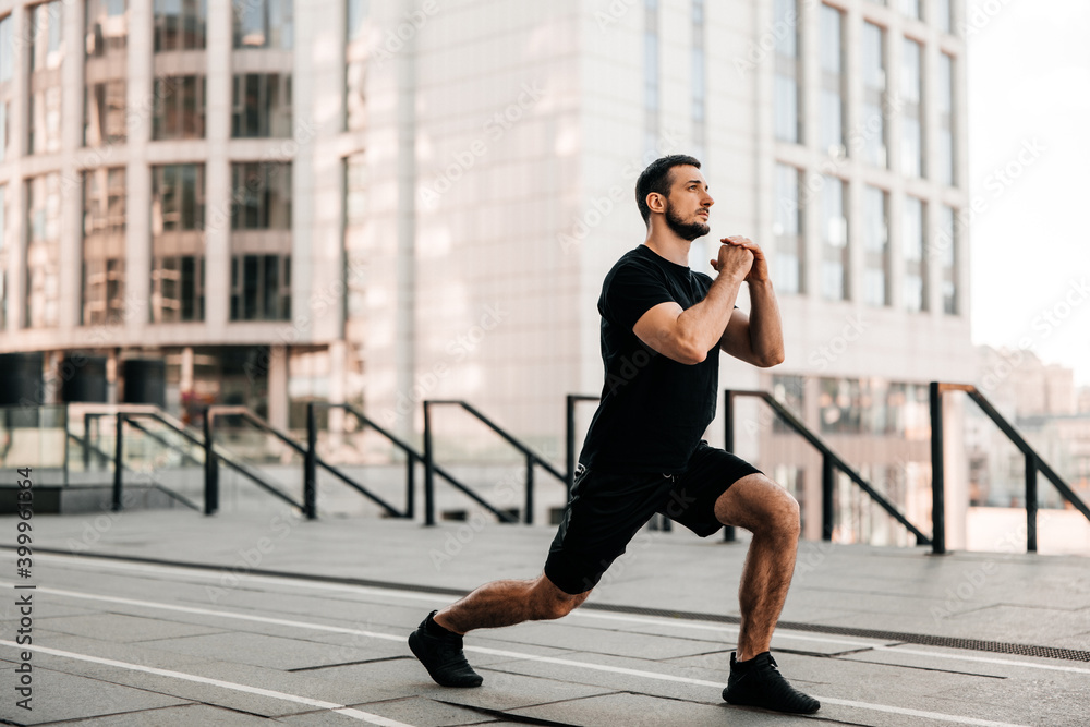Fit man doing lunge forward. Sport in big city concept. Strong man stretching legs before jogging. Healthcare concept. Runner in black sportswear. Urban sport concept. Fit man doing lunge forward. Sport in big city concept. Strong man stretching legs before jogging. Healthcare concept. Runner in black sportswear. Urban sport concept.