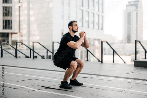 Fit man training air squat exercise. Fit male exercising crossfit outside. Young handsome caucasian male fitness model and instructor outdoors. Black sportswear. Sport in big city concept.