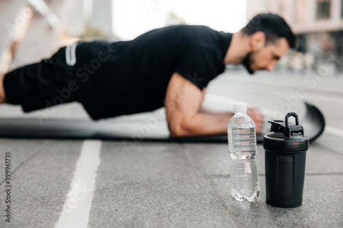 Concentrated man doing plank position while exercising on black yoga mat. Water bottles lying near. Blurred man in black sportswear exercising outdoors. Morning warm up. Side view.