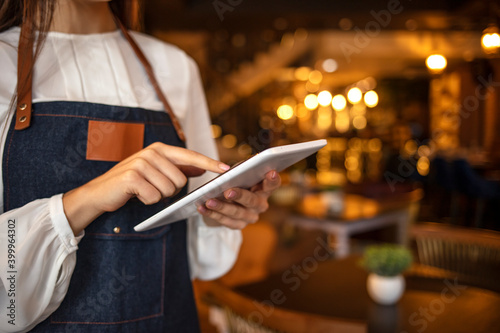 Close-up of female owner standing in her cafe using digital tablet. Female barista standing in coffee shop using digital tablet. Young female owner using digital tablet while standing in cafe