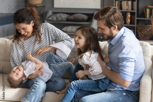 Overjoyed young Caucasian family sit on sofa in living room play tickle little daughters. Happy mother and father feel playful have fun with two small girls children, enjoy weekend at home together.