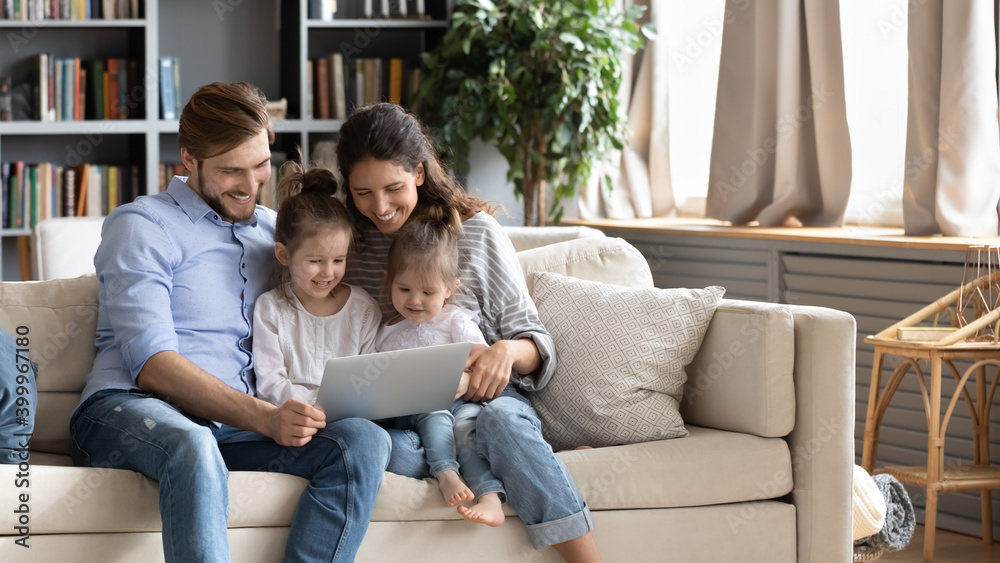 Wide banner panoramic view of happy young Caucasian family renters with two small daughters relax on sofa using laptop together. Smiling parents with little girls children browse internet on computer. Wide banner panoramic view of happy young Caucasian family renters with two small daughters relax on sofa using laptop together. Smiling parents with little girls children browse internet on computer.
