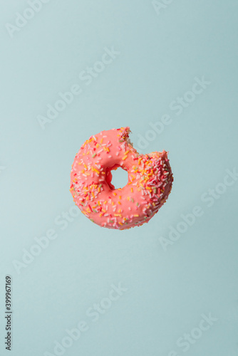 Pink donut with colorful sprinkles with a bite flying on blue background.