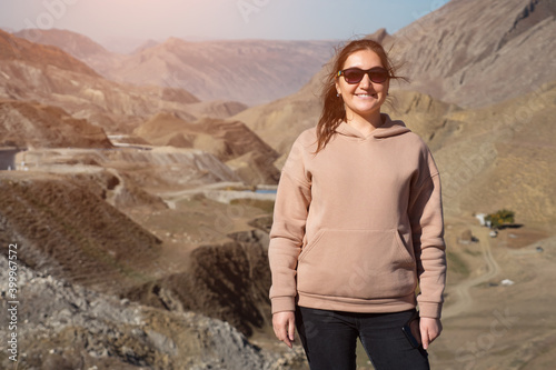 Happy brunette woman with elegant sunglasses in hoodie looks at camera and enjoys wonderful landscape of hilly valley with road in highland on sunny day