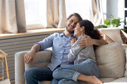 Happy millennial Caucasian couple sit relax on comfortable couch at home hugging and cuddling. Smiling young man and woman rest on sofa in living room enjoy family romantic leisure weekend together.